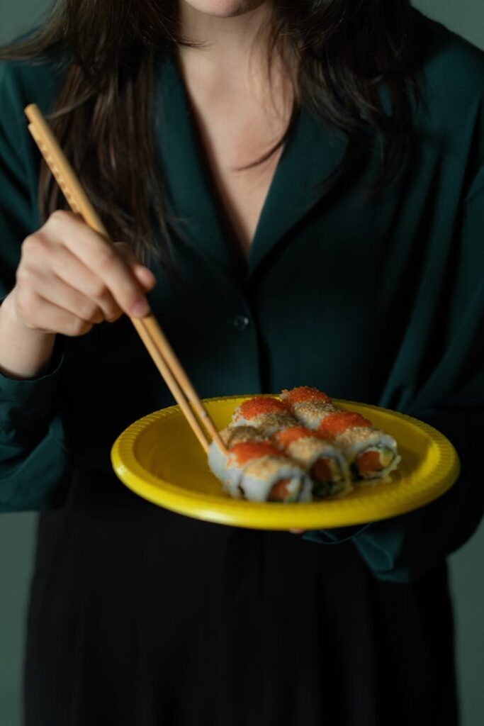 Close-up of a woman holding sushi on a yellow plate with chopsticks, showcasing Asian cuisine.